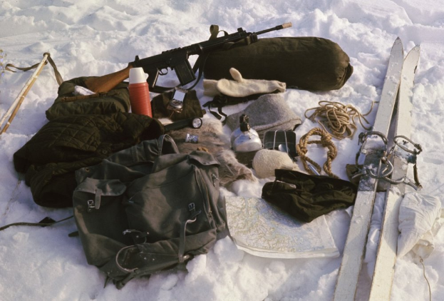 Ein schneebedeckter Boden mit verstreuten Gegenständen, darunter eine Tasche, eine Waffe, eine Flasche, Handschuhe, ein Paar Skier, ein Seil und andere Gegenstände.