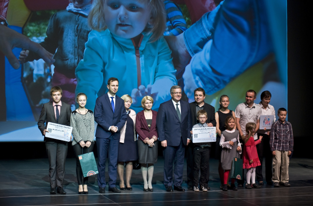 Eine Gruppe von Menschen auf einer Bühne, einige halten Taschen und Urkunden in den Händen, im Hintergrund ein Bildschirm mit einem Bild von einem Kind und anderen Menschen.