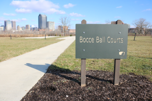 Ein verwittertes Schild am Boden in der Nähe einer grasigen Fläche trägt die Aufschrift "bocce ball courts in Indianapolis, Indiana" mit Bäumen, einem Zaun, Gebäuden und einem bewölkten Himmel im Hintergrund.