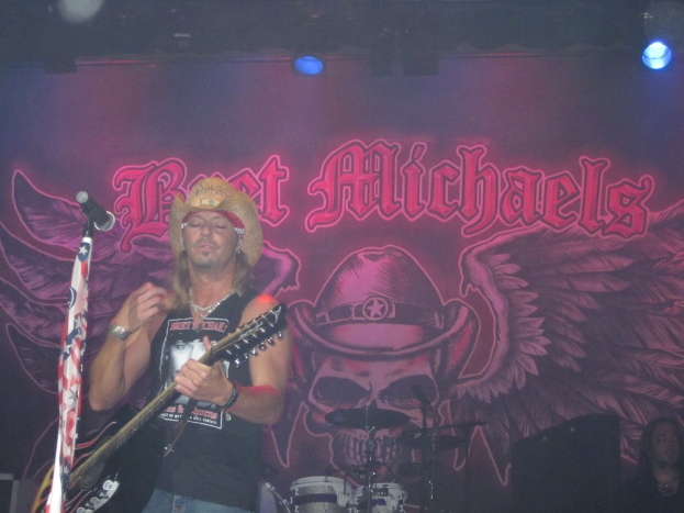 Eine Musik-Konzert-Szene mit einem Gitarristen, der auf der Bühne spielt, einer Person, die in der unteren rechten Ecke sitzt, einem Banner im Hintergrund und Drogen, die in der Mitte unten platziert sind.