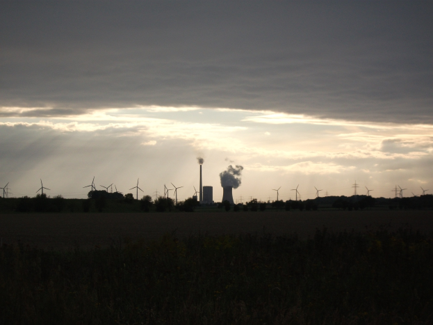 Kraftwerk mit Windrädern vor einem bewölkten Himmel, umgeben von Grün am Boden.