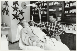 Ein Mann lächelt im Zahnarztstuhl, während ein anderer Mann neben ihm steht und ein Tablett hält, mit einem Waschbecken, Sternen, einer Uhr und Regalen voller Flaschen im Hintergrund.