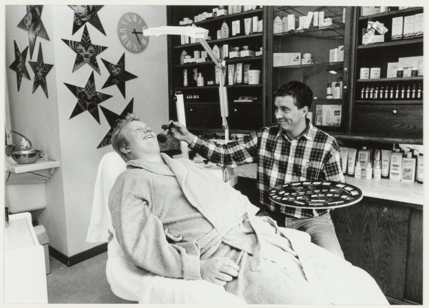Ein Mann lächelt im Zahnarztstuhl, während ein anderer Mann neben ihm steht und ein Tablett hält, mit einem Waschbecken, Sternen, einer Uhr und Regalen voller Flaschen im Hintergrund.