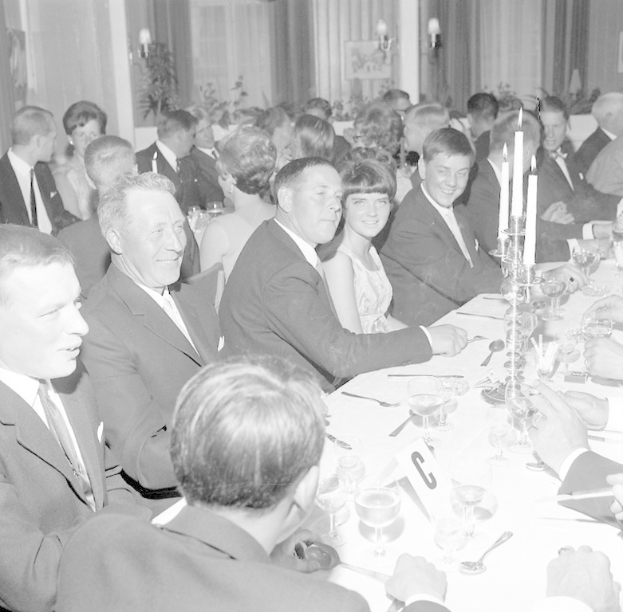 Eine Gruppe von Menschen sitzt um einen langen Tisch, lächelt und genießt die Gesellschaft des anderen, mit Geschirr wie Gläsern, Löffeln, Gabeln, Messern und Kerzenhaltern, vor einem Hintergrund aus Vorhängen und Wandlampen.