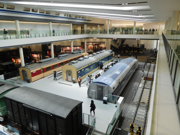 Ein großer Museumsraum mit verschiedenen Zügen in unterschiedlichen Größen und Farben, mit Menschen, die die Ausstellungsstücke beobachten und Deckenlampen, die den Raum beleuchten.