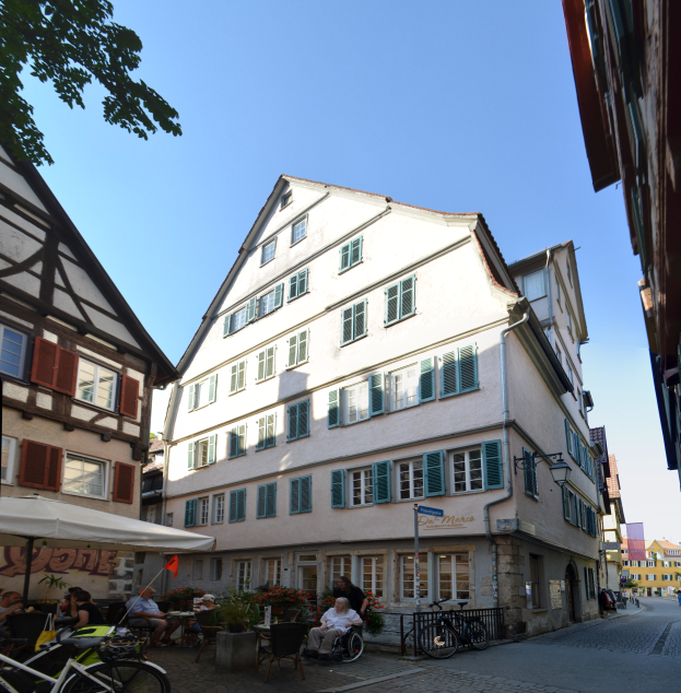 Ein Kopfsteinpflasterweg in Rothenburg ob der Tauber, Deutschland, mit Menschen auf Stühlen, parkenden Fahrrädern, blühenden Pflanzen, einem Zelt, einem Schild, einem Straßenpfahl, einer Gruppe von Gebäuden mit Fenstern, einem Baum und einem bewölkten Himmel.