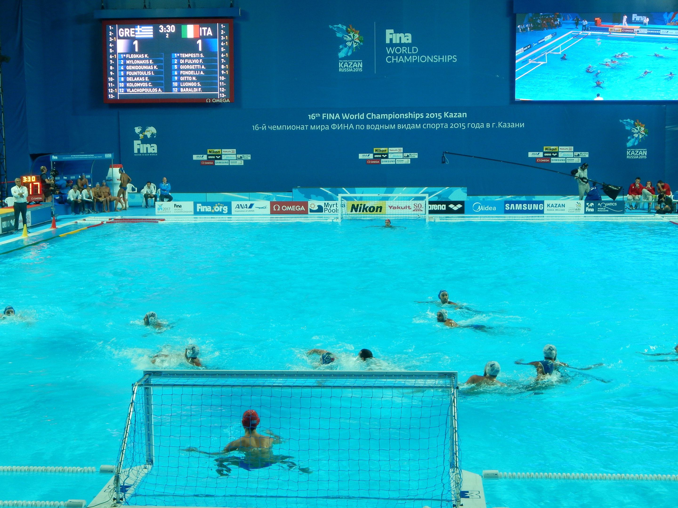 Gruppe von Athleten, die Wasserball in einem Pool während der FINA-Weltmeisterschaften 2016 in Kazan spielen, mit Zuschauern links und einer Anzeigetafel im Hintergrund.