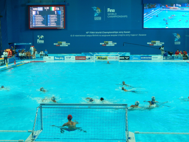 Gruppe von Athleten, die Wasserball in einem Pool während der FINA-Weltmeisterschaften 2016 in Kazan spielen, mit Zuschauern links und einer Anzeigetafel im Hintergrund.