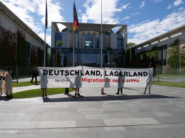 Eine Gruppe von Menschen hält ein Transparent mit der Aufschrift "Deutschland, Lagerland Migration ist kein Verbrechen" vor einem Gebäude mit Fenstern, umgeben von Gras, einem Metallzaun, Pflanzen, Bäumen und Fahnen auf Ständen, unter einem bewölktem Himmel.