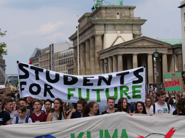 Gruppe von Schülern marschiert in Berlin mit einem farbenfrohen "Students for Future"-Schild gegen eine Kulisse aus Gebäuden, Bäumen und Himmel.