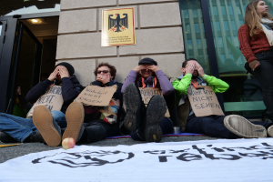 Eine Gruppe von Menschen sitzt vor einem Gebäude auf dem Boden und hält Schilder, mit einem Banner mit Text und einem Apfel auf der Straße, und eine Frau steht rechts daneben und sieht besorgt aus.
