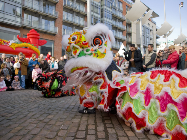 Ein farbenfrohes chinesisches Neujahrsfest in Amsterdam mit einer Löwen-Tanzvorstellung vor einer Zuschauermenge, einige halten Kameras, vor dem Hintergrund von Gebäuden, Laternenmasten und einem klaren blauen Himmel.