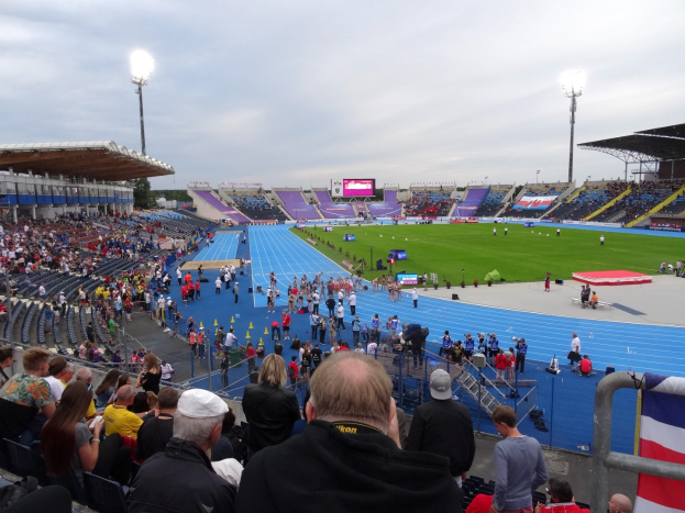 Ein großes Stadion voller Zuschauer bei einem Fussballspiel, mit Menschen auf Sitzen und Stehplätzen, Geländern, einer Fahne, Flutlicht, einem Bildschirm, Bäumen und einem bewölkten Himmel im Hintergrund.