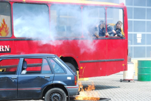 Ein roter Doppeldeckerbus mit Rauch, der aus ihm austritt, mit drei sichtbaren Passagieren, neben einem Auto geparkt, vor einem Gebäude mit Glasfenstern und einem Fass auf der rechten Seite.