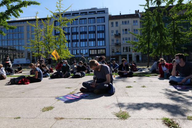Eine Gruppe von Menschen sitzt vor einem Gebäude in Berlin auf dem Boden, einige tragen Masken, mit verstreuten Taschen und Gegenständen, unter einem klaren blauen Himmel.