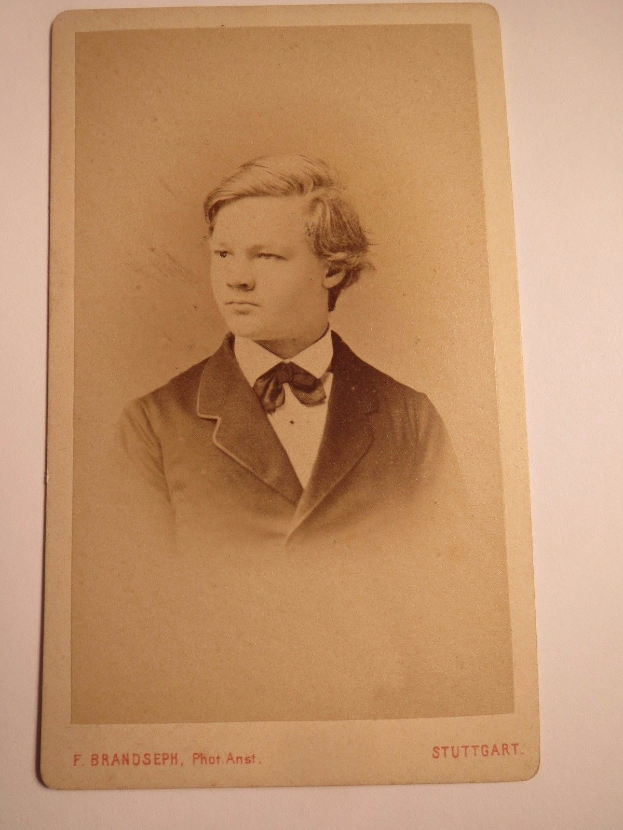 Schwarzes und weißes Foto eines jungen Mannes in Anzug und Schleife, identifiziert als F. Brandseph Stuttgart, aufgenommen in den frühen 1900er Jahren, befestigt an einer weißen Oberfläche mit Text unten.