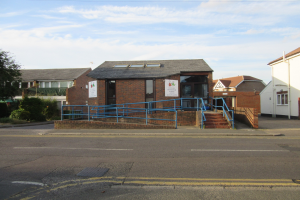 Ein Schulgebäude mit einem blauen Geländer, umgeben von Häusern, Stufen, Geländern, Texttafeln, Pflanzen, Bäumen und einem bewölkten Himmel.