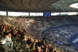 Große Zuschauermenge in einem Stadion bei einem Fußballspiel, mit einer Bühne rechts, Fahnen, Stangen, einem Bildschirm und der Allianz Arena in München, Deutschland im Hintergrund.