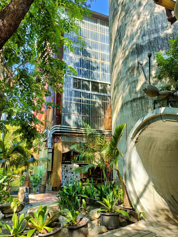 Ein Innenhof mit Topfpflanzen und Bäumen vor einem modernen, umweltfreundlichen Hotelgebäude, umgeben von üppiger Vegetation unter einem klaren blauen Himmel.