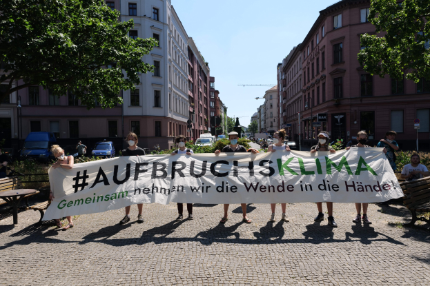 Eine Gruppe von Menschen in Masken und mit einem Banner mit der Aufschrift 'Aufbruchsklima' protestiert gegen den Klimawandel in Berlin, Deutschland, vor einem Gebäude, das von Bänken, Tischen, Pflanzen, Bäumen, Fahrzeugen, Laternenmasten und Schildern umgeben ist.
