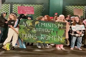Eine Gruppe von Menschen mit einem Banner mit der Aufschrift "Kein Feminismus ohne Trans-Frauen" und verschiedenen Schildern, die auf einem öffentlichen Platz mit einer Wand und einer Rolllade im Hintergrund stehen.