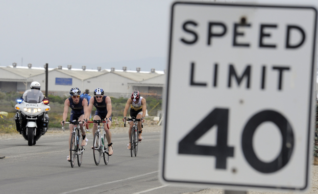 Gruppe von Radfahrern, die eine Straße mit einem Tempolimit-Schild hinunterfahren, Gebäude und Bäume im Hintergrund unter einem klaren blauen Himmel.