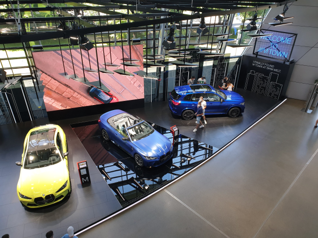 Eine Gruppe von Menschen, die im BMW Museum in München stehen, mit Autos zur Linken und einem Schild mit der Aufschrift "BMW Museum München" zur Rechten, mit Blick auf Bäume und Gebäude draußen.