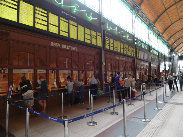Menschen in einer Warteschlange an einem Bahnhofsschalter mit Stangen, Bändern, Informationsschildern, Deckenleuchten und Treppen mit Gelöndern im Hintergrund.