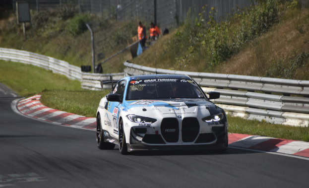 Ein BMW M8 GT3 Rennauto auf einer Rennstrecke, umgeben von einem Zaun, Gras, Pflanzen und einer Gruppe von Menschen, mit Text auf dem Auto und einem leicht unscharfen Hintergrund.