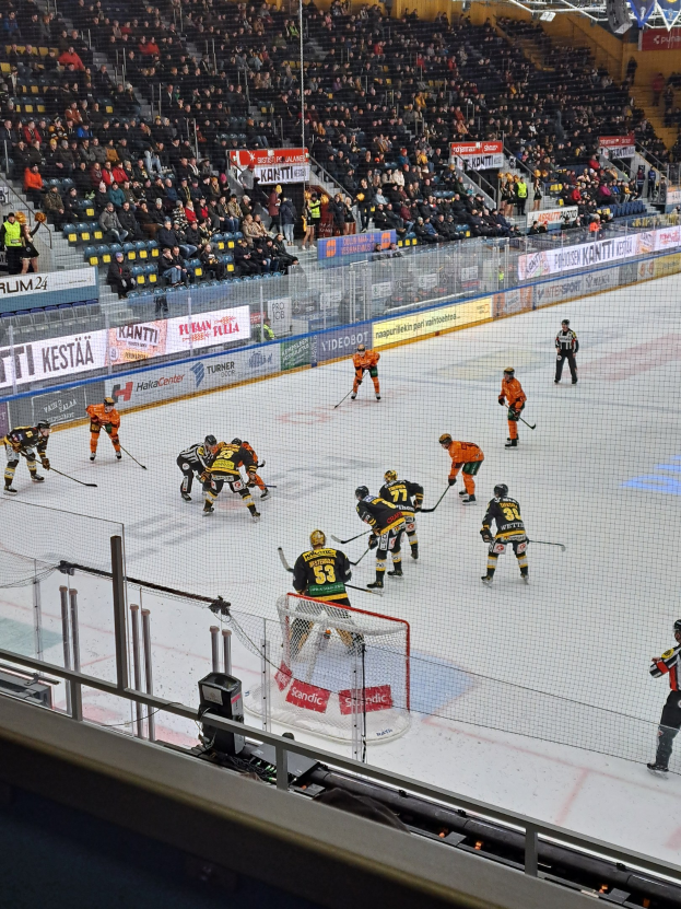 Eine Gruppe von Menschen, die Hockey auf einem Eisring mit einem Netz und einer umgebenden Absperrung spielen, Zuschauern im Hintergrund und Bannern mit Text, unter hellen Stadionlichtern.