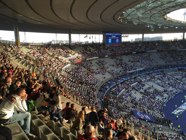 Große Menschenmenge in einem Stadion bei einem Fußballspiel, mit einer Bühne auf der rechten Seite, Fahnen, Stangen, einem Bildschirm und der Allianz Arena in München, Deutschland im Hintergrund.
