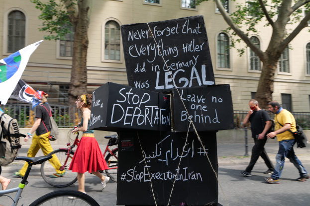 Eine Gruppe von Menschen, die mit Fahrrädern die Straße entlanggehen, einige tragen Taschen und einer hält eine Fahne, im Hintergrund sind Bäume, ein Gebäude mit Fenstern und ein Schild mit der Aufschrift "Stop Deportation" zu sehen.