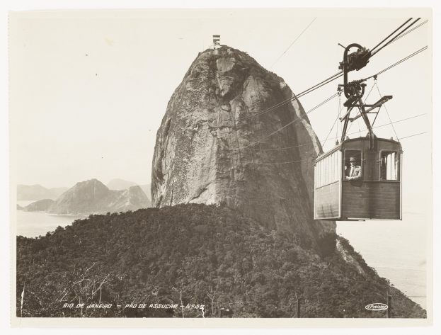 Schwarz-weiß-Foto einer Seilbahn, die einen Berg hinauffährt, mit einer Person darin, umgeben von Bäumen, Hügeln und Wasser; unten ist Text zu sehen.