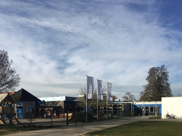 Großes Gebäude mit einem grasigen Bereich davor, umgeben von einem Metallzaun, Bäumen, Schildern, einem Fahrrad, nahegelegenen Häusern und einem bewölkten Himmel, identifiziert als der neue Spielbereich für Kinder in der Schule.