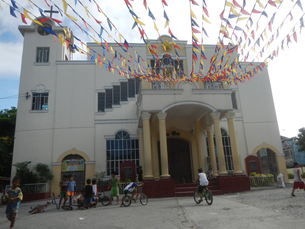 Gruppe von Menschen, die vor einer weißen Kirche mit architektonischen Details Fahrräder fahren, umgeben von Grün, einem Zaun, einer Fahne, Bäumen und bunten Flaggen, die an einem Seil befestigt sind, unter einem bewölkten Himmel.