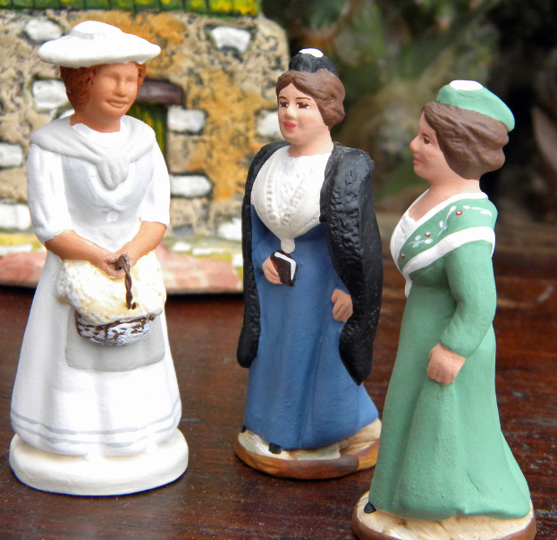 Drei geschnitzte Puppen sind auf einer Holzplatte mit Spielzeughäusern im Hintergrund zu sehen.