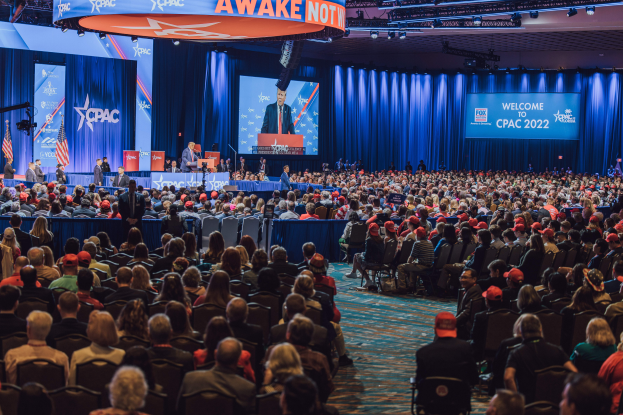 Eine große Menschenmenge auf Stühlen sitzend, die einer Bühne mit einigen Menschen darauf zugewandt ist, blaue Vorhänge und ein Bildschirm im Hintergrund und eine orangefarbene Tafel oben, die das CPAC 2020-Event am 25. Oktober anzeigt.