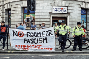 Eine Gruppe von Menschen mit einem "Kein Platz für Faschismus"-Schild auf einem Fußweg, mit zwei Polizeibeamten davor, Fahrrädern, einer Ampel und einem Gebäude im Hintergrund.