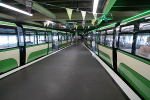 Ein grüner und weißer Zug mit Passagieren fährt auf Schienen neben einem Bahnsteig in einem Münchner Bahnhof, mit einer Decke, die mit Lichtern und dekorativen Flaggen geschmückt ist.