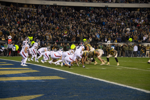 Ein Fußballspiel im Gange mit Spielern auf dem Boden in Sportkleidung und Helmen, aus der Perspektive der Tribüne betrachtet.