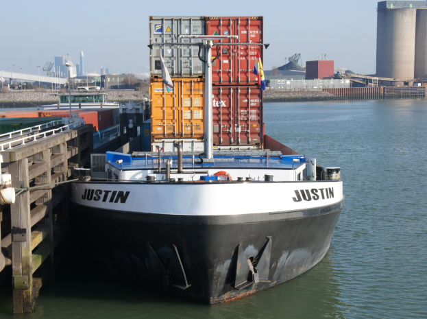 Großer Containerfrachter liegt neben einem Kai, mit einer Brücke links, Gebäuden, Bäumen und Himmel im Hintergrund.