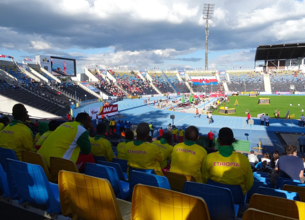 Eine Gruppe von Menschen in einem vollen Stadion bei einem Fußballspiel mit Schildern, einem Bildschirm und einem bewölkten Himmel im Hintergrund.