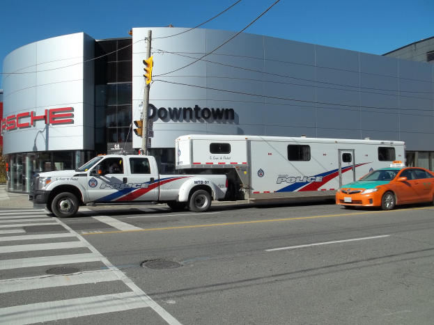 Ein Polizeiwagen vor einem Gebäude mit Text, eine Ampel mit Stange und Drähten im Vordergrund und Bäume mit einem klaren blauen Himmel im Hintergrund.