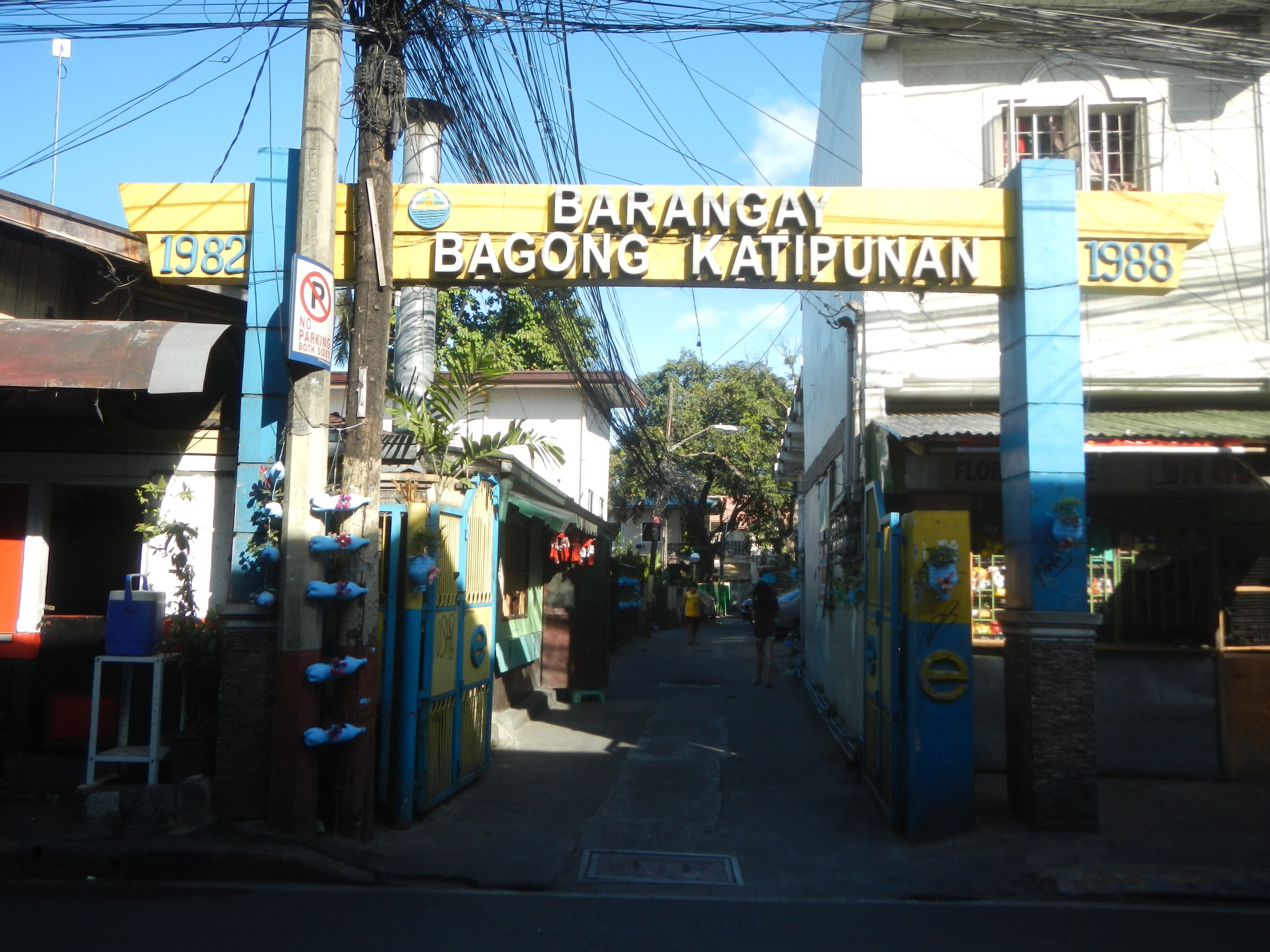 Eingangsbogen zum Barangay Bagong Katipunan, Philippinen, mit Gebäuden, Strommasten, Schildern und Passanten unter einem bewölkten Himmel.