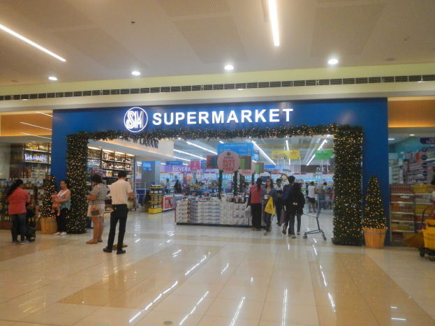 Menschen vor einem Supermarkt mit Einkaufstaschen, einem Einkaufswagen, Topfpflanzen, Regalen, Schildern und einer Decke mit Deckenleuchten.