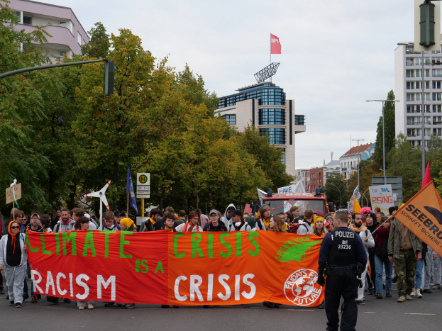 Eine Gruppe von Menschen marschiert eine von Bäumen gesäumte Straße entlang und hält ein Banner mit der Aufschrift "Klimakrise ist eine Krise", mit Gebäuden und einem klaren blauen Himmel im Hintergrund.