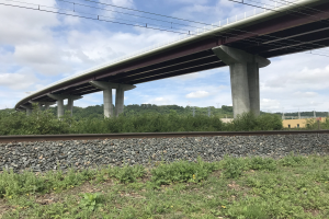Eisenbahnschiene mit wolkenverhangenem Himmel, eine Brücke in Renovierung, ein Gebäude im Hintergrund, umgeben von Steinen, Gras, Pflanzen, Bäumen, Pfählen und Drähten.