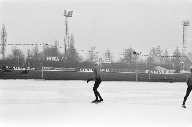 Zwei Menschen, die auf einer Eisbahn im Schnee Schlittschuhlaufen, umgeben von einem Zaun, Bannern, Pfosten, Lichtern, Bäumen und einem klaren Himmel, dargestellt in Schwarz-Weiß.