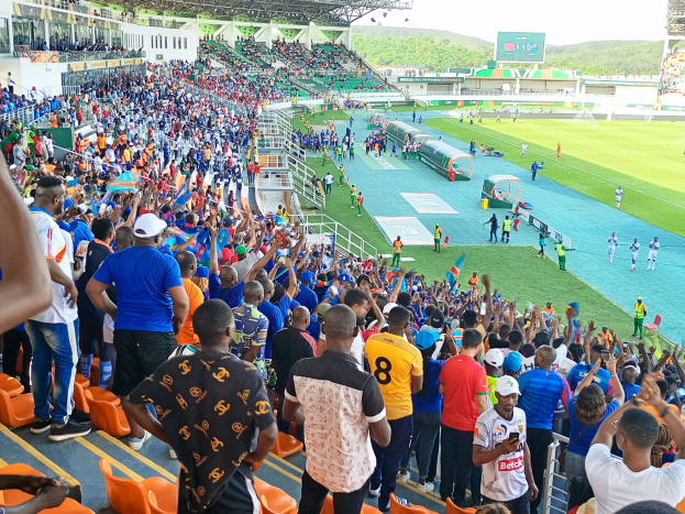 Eine große Menschenmenge versammelt sich vor einem eingezäunten Fußballfeld mit Hügeln und einem blauen Himmel im Hintergrund und erwartet das Spiel der 2019 African Cup of Nations.