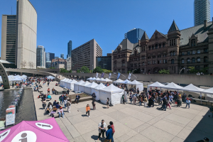 Eine große Menschenmenge versammelt sich vor Zelten vor einer Toronto-Skyline, mit einer Brücke links, Wasser rechts und Bäumen und Gebäuden im Hintergrund, unter einem sichtbaren Himmel.